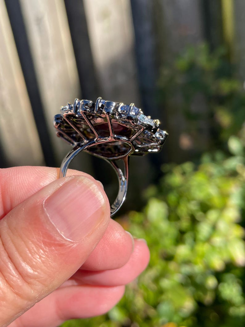 May include: A silver ring with a large, ornate setting. The setting is a cluster of small, dark stones surrounding larger, clear stones. The ring is held up by a person's fingers, with a blurred green background.
