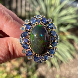 May include: An oval-shaped ring featuring a large, dark opal with iridescent flashes of color. The opal is surrounded by a halo of blue sapphires and smaller, light blue gemstones, set in a gold band. The ring is a statement piece.