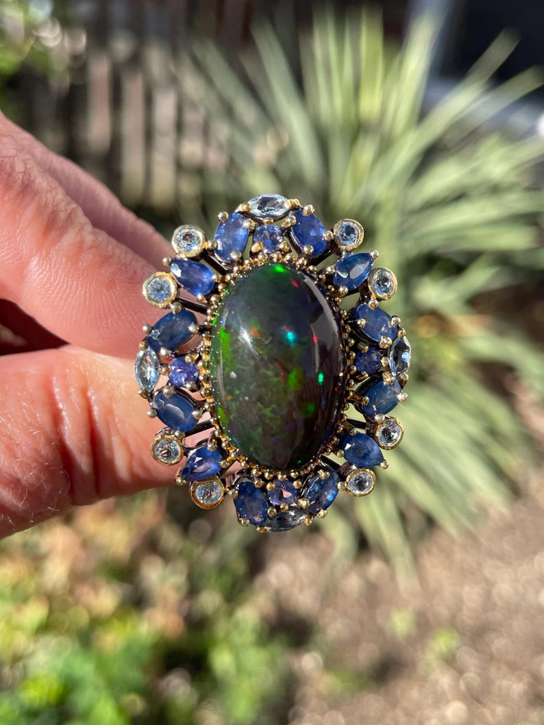 May include: An oval-shaped ring featuring a large, dark opal with iridescent flashes of color. The opal is surrounded by a halo of blue sapphires and smaller, light blue gemstones, set in a gold band. The ring is a statement piece.