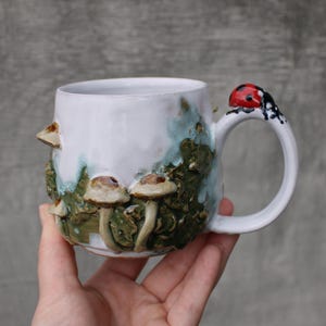May include: A white ceramic mug featuring a textured, sculpted forest scene with raised brown mushrooms and green foliage. The handle is adorned with a small, painted red and black ladybird perched at the top, adding a distinct detail to the piece.
