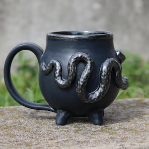 Peut inclure: Une tasse en céramique noir mat avec un corps arrondi et trois pieds courts. La tasse présente un serpent en relief à la finition métallique texturée enroulé autour de la surface extérieure. Elle possède une anse incurvée et un bord sombre.