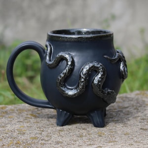 Peut inclure: Une tasse en céramique noir mat avec une forme arrondie rappelant un chaudron, dotée de trois petits pieds à la base et d'une anse incurvée. Un relief de serpent noir brillant et texturé enveloppe l'extérieur de la tasse.