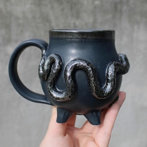 Peut inclure: Une tasse en céramique gris foncé mat avec un corps arrondi, une anse unique et trois pieds courts. L'extérieur présente un serpent en relief texturé à l'aspect métallique qui s'enroule autour du centre, avec un bord sombre et brillant.