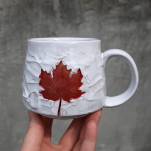 Peut inclure: Une tasse en céramique blanche avec une surface texturée et irrégulière, présentant une impression unique et proéminente de feuille d'érable rouge foncé au centre. La tasse possède un corps arrondi et une anse incurvée sur le côté droit.