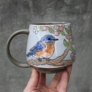 May include: A ceramic mug with a light grey base and a dark grey handle. The mug features a hand-painted illustration of a blue bird with an orange chest perched on a brown tree branch with green leaves and small pink flowers against a white background.
