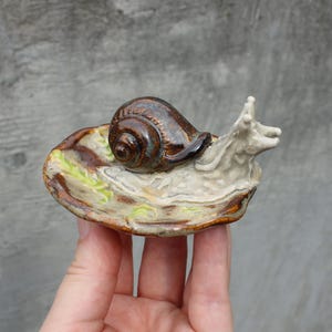 Könnte beinhalten: Eine Keramikfigur einer Schnecke mit braunem und beigem Gehäuse und cremefarbenem Körper. Die Schnecke ruht auf einer blattförmigen Basis mit grünen und braunen Akzenten. Die Figur wird in einer Hand gehalten.