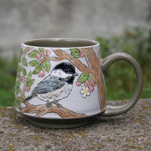 May include: A ceramic mug with a white base and a grey handle and base. The mug features a hand-painted illustration of a black-capped chickadee perched on a brown tree branch with green leaves and small pink and white flowers surrounding it.