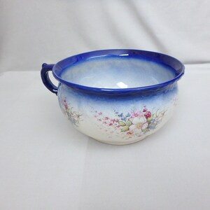 May include: A ceramic chamber pot featuring a deep blue rim and handle, transitioning into a white body. The exterior is decorated with a delicate floral pattern in shades of pink, green, and blue, showcasing a classic rounded basin shape.