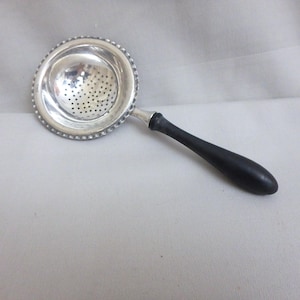 May include: A silver-colored tea strainer featuring a circular bowl with a perforated base and a decorative beaded rim. The strainer is attached to a smooth, solid black handle that extends outward, providing a contrast to the metallic finish.