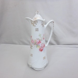 May include: A tall white porcelain coffee pot featuring a delicate floral pattern of pink roses and green leaves. The vessel includes a matching lid with a pointed finial, a curved handle, and gold filigree accents along the rim and base.