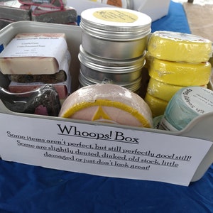 May include: A grey plastic bin filled with various bath and body products, including soaps, bath bombs, and tin containers. A sign on the front of the bin reads "Whoops! Box Some items aren't perfect, but still perfectly good still! Some are slightly dented, dinked, old stock, little damaged or just don't look great!"