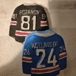 K&ouml;nnte beinhalten: Zwei dekorative Hockey-Trikot-Ornamente. Eines ist schwarz mit dem Namen "ROZANOV" und der Nummer "81" in Wei&szlig;. Das andere ist blau mit dem Namen "HOLLANDER" und der Nummer "24" in Wei&szlig;. Beide haben rot-wei&szlig;e Streifen.