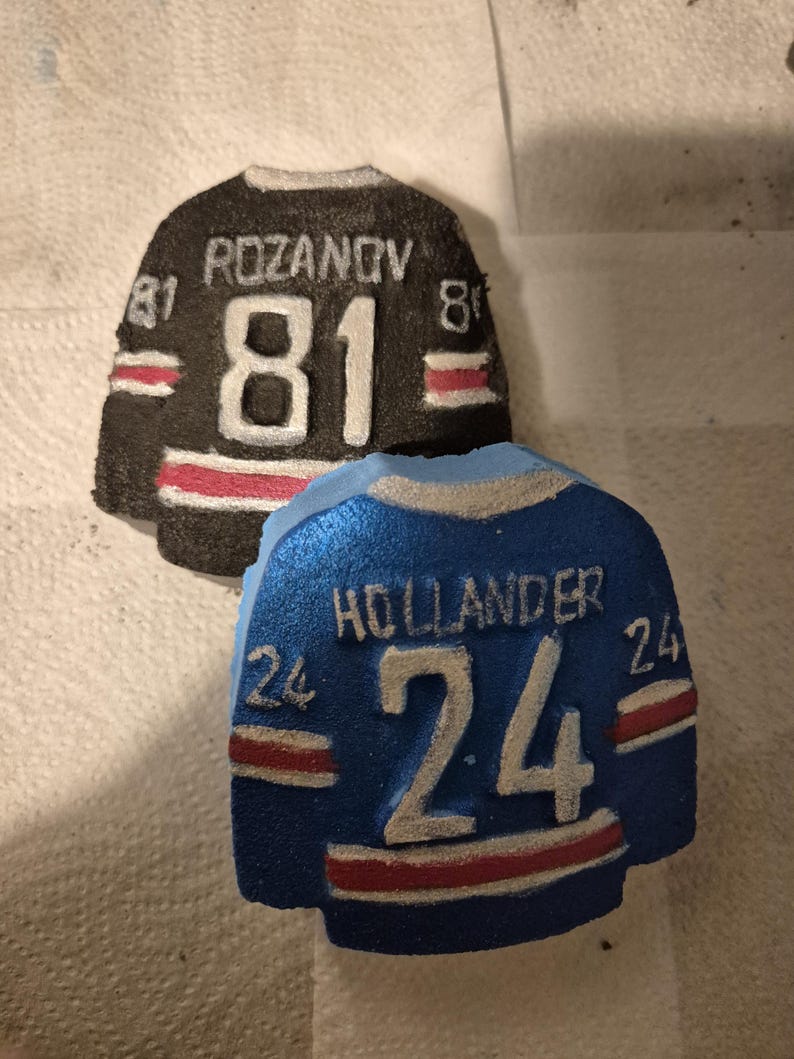 K&ouml;nnte beinhalten: Zwei dekorative Hockey-Trikot-Ornamente. Eines ist schwarz mit dem Namen "ROZANOV" und der Nummer "81" in Wei&szlig;. Das andere ist blau mit dem Namen "HOLLANDER" und der Nummer "24" in Wei&szlig;. Beide haben rot-wei&szlig;e Streifen.