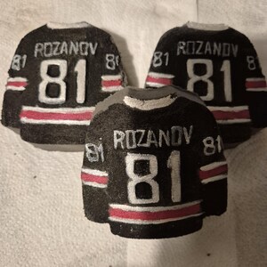 K&ouml;nnte beinhalten: Drei schwarze, hockey-Trikot-f&ouml;rmige Objekte mit dem Namen "ROZANOV" und der Nummer "81" in Wei&szlig;. Jedes Trikot hat einen roten Streifen am unteren Rand und wei&szlig;e Akzente. Die Trikots sind auf einer wei&szlig;en Oberfl&auml;che angeordnet.
