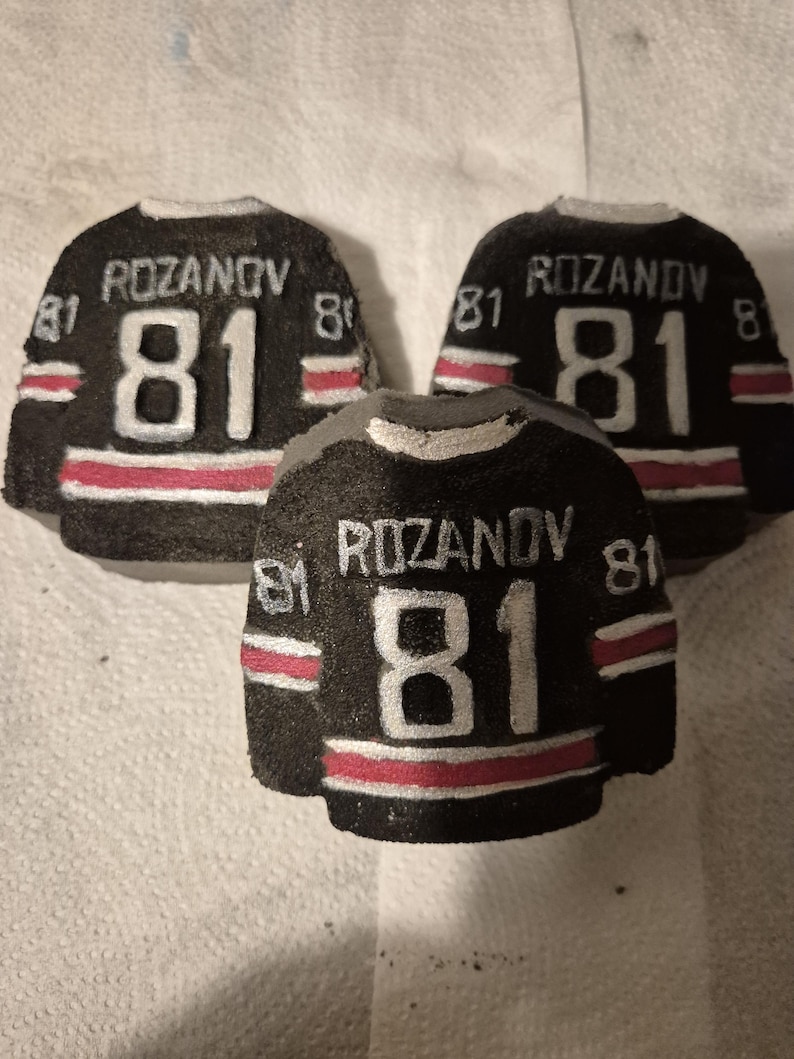 K&ouml;nnte beinhalten: Drei schwarze, hockey-Trikot-f&ouml;rmige Objekte mit dem Namen "ROZANOV" und der Nummer "81" in Wei&szlig;. Jedes Trikot hat einen roten Streifen am unteren Rand und wei&szlig;e Akzente. Die Trikots sind auf einer wei&szlig;en Oberfl&auml;che angeordnet.