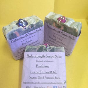 Puede incluir: Tres barras de jabón morado y verde con un dado de 13 caras incrustado en cada una. El jabón está etiquetado como "Helensburgh Soapy Suds" y "Fun Scary! Laudna (Critical Role) Dragons Blood Processed Soap". El jabón es vegano, libre de crueldad y de origen sostenible. El jabón pesa 100 g.