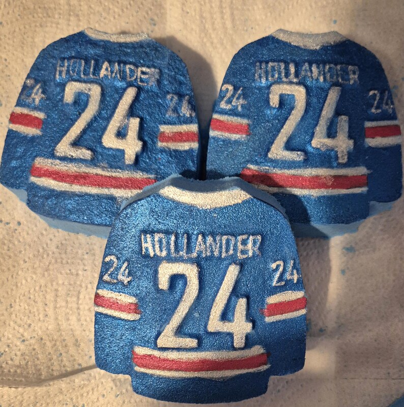 K&ouml;nnte beinhalten: Drei blaue Hockey-Trikot-f&ouml;rmige Objekte mit dem Namen "HOLLANDER" und der Nummer "24" in Wei&szlig;. Jedes Trikot hat rote und wei&szlig;e Streifen am unteren Rand. Die Trikots sind in einem dreieckigen Muster angeordnet.