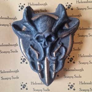 Puede incluir: Una bomba de baño negra y dorada con forma de calavera con cuernos. La bomba de baño está sobre un fondo marrón con el texto "Helensburgh Soapy Suds" repetido.