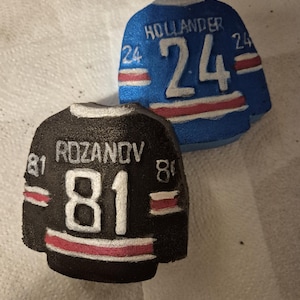 K&ouml;nnte beinhalten: Zwei Miniatur-Hockeytrikots, eines in Blau und eines in Schwarz, werden gezeigt. Das blaue Trikot tr&auml;gt den Namen "HOLLANDER" und die Nummer "24". Das schwarze Trikot zeigt den Namen "ROZANOV" und die Nummer "81". Beide Trikots haben wei&szlig;e und rote Akzente.