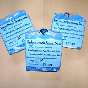 Peut inclure: Trois savons artisanaux avec des tourbillons bleus et blancs, chacun enveloppé d'une étiquette indiquant "Helensburgh Soapy Suds". Les étiquettes indiquent les ingrédients et le texte "Ever Bright, Ever Right! (Critical Role)". Chaque savon pèse 100g.