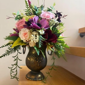 May include: A floral arrangement in a dark bronze-toned metal urn with two side handles. The bouquet features a mix of pink, purple, and deep burgundy flowers, including roses and anemones, accented with various green ferns and foliage.