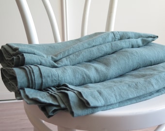 May include: A folded teal linen sheet on a white chair. The sheet is made of natural linen fabric and has a soft, textured surface.
