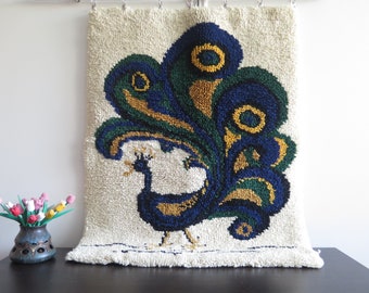 Op de afbeelding: Een wit wandtapijt met een blauw, groen en gouden pauwontwerp. De pauw is afgebeeld met zijn staartveren uitgespreid.