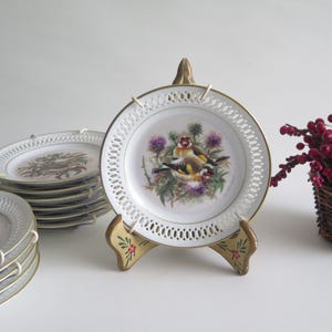 Könnte beinhalten: Eine Sammlung dekorativer Porzellanteller mit goldenen Rändern und aufwendigen Lochmustern. Jeder Teller zeigt eine detaillierte Vogelillustration mit floralem Hintergrund. Ein Teller wird auf einem dekorativen Ständer präsentiert.
