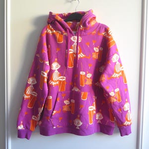 Könnte beinhalten: Ein leuchtend magentafarbener Kapuzenpullover mit einem sich wiederholenden orangefarbenen Grafikdruck einer Zeichentrickfigur mit Dutt und hoher Brille. Der langärmelige Pullover hat eine Kapuze mit Kordelzug, gerippte Bündchen und eine Kängurutasche.