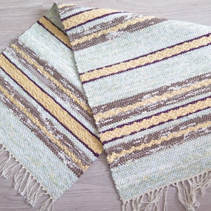 May include: A woven rug with a light blue background and stripes of brown, yellow, and purple. The rug has a fringe on all sides.