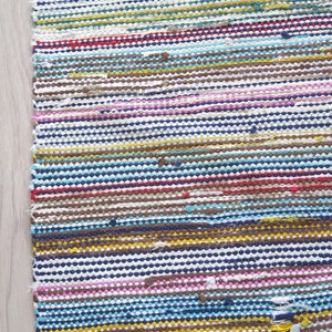 May include: A colorful striped woven rug with a fringe edge. The rug is made of recycled materials and has a variety of colors, including blue, yellow, pink, brown, and white.