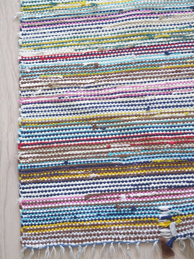 May include: A colorful striped woven rug with a fringe edge. The rug is made of recycled materials and has a variety of colors, including blue, yellow, pink, brown, and white.