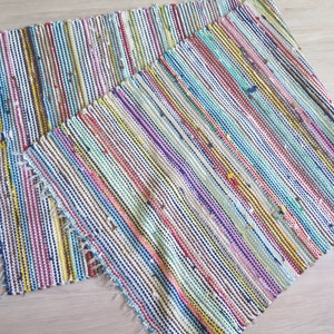 May include: Two colorful striped woven rugs with frayed edges. The rugs are made of recycled materials and have a variety of colors, including blue, green, red, yellow, and white.