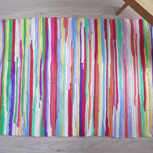May include: A rectangular, handwoven rug with vertical stripes in a variety of colors including red, yellow, green, purple, and blue. The rug has a fringed edge and is placed on a light-colored wooden floor. The rug is a home decor item.