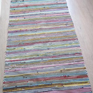May include: A colorful striped rug with a variety of colors, including red, blue, yellow, green, and pink. The rug is made of woven fabric and has a fringed edge.