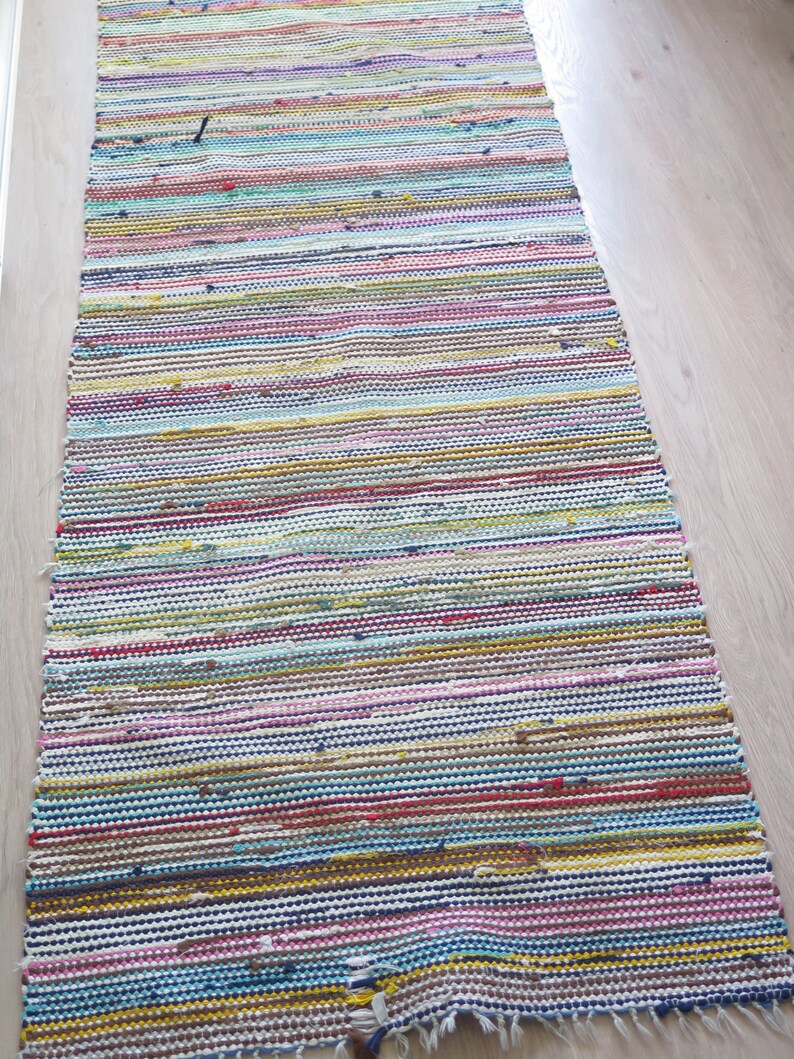 May include: A colorful striped rug with a variety of colors, including red, blue, yellow, green, and pink. The rug is made of woven fabric and has a fringed edge.
