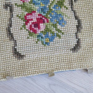 May include: A close-up of a beige needlepoint cushion with a floral design featuring pink, blue, and green flowers. The cushion has a brown border and is trimmed with beige fabric.