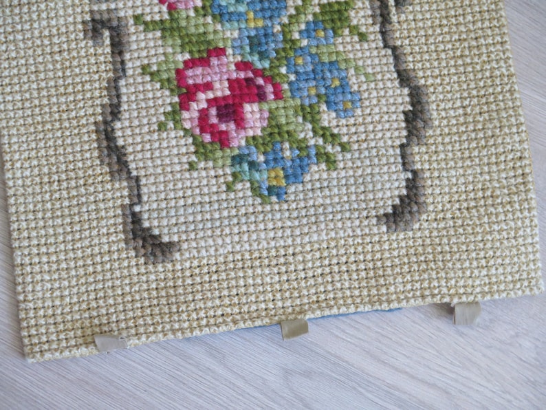 May include: A close-up of a beige needlepoint cushion with a floral design featuring pink, blue, and green flowers. The cushion has a brown border and is trimmed with beige fabric.