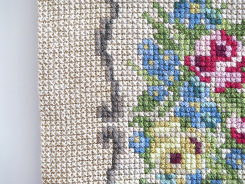 May include: Close-up of a beige cross-stitch fabric with a floral design. The design features blue, green, yellow, pink, and red flowers.
