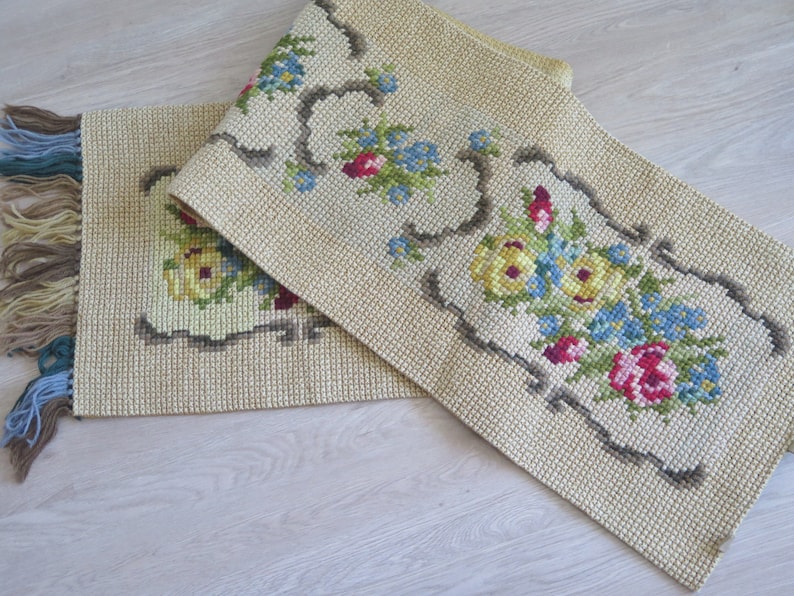 May include: Two beige embroidered table runners with floral designs. The runners have a fringe border and feature yellow, blue, red, and green flowers.