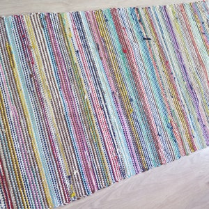 May include: A colorful woven rug with stripes of red, blue, yellow, green, pink, and brown. The rug is made of recycled materials and has a natural, rustic look.