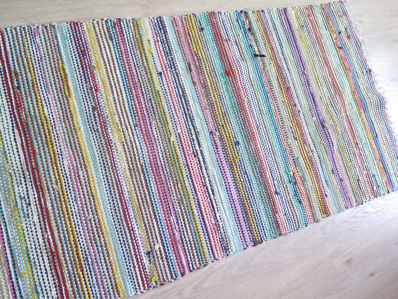 May include: A colorful woven rug with stripes of red, blue, yellow, green, pink, and brown. The rug is made of recycled materials and has a natural, rustic look.