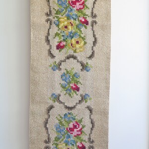 May include: A beige fabric wall hanging with three embroidered floral designs. Each design features a cluster of pink, yellow, and blue flowers with green leaves, framed by a brown border.