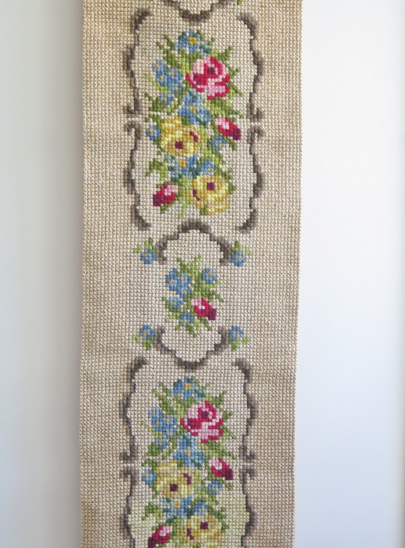 May include: A beige fabric wall hanging with three embroidered floral designs. Each design features a cluster of pink, yellow, and blue flowers with green leaves, framed by a brown border.