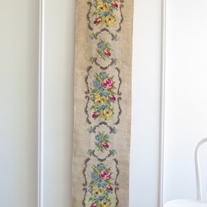 May include: A beige tapestry with a floral design featuring red, yellow, and blue flowers. The tapestry has a fringe border and is hanging on a white wall.