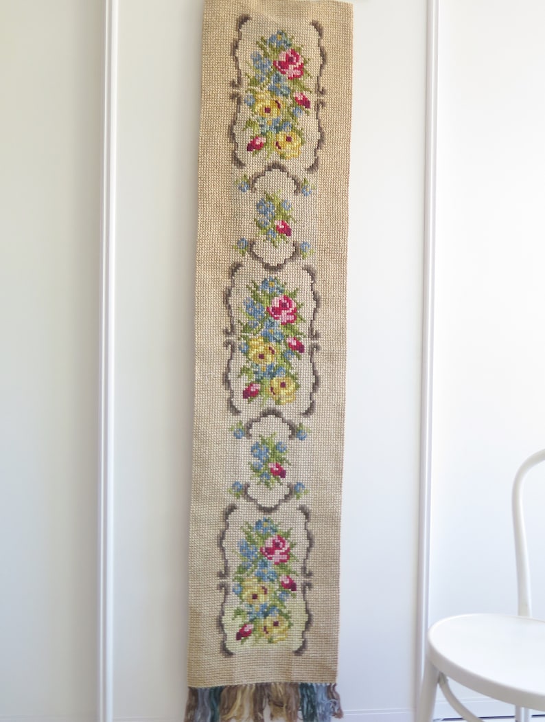 May include: A beige tapestry with a floral design featuring red, yellow, and blue flowers. The tapestry has a fringe border and is hanging on a white wall.