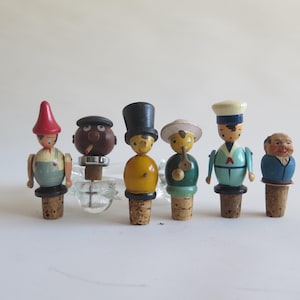 Può includere: Una collezione di sei tappi per bottiglie vintage in legno dipinti a mano. Ogni tappo presenta un design unico, tra cui uno gnomo, un uomo con la pipa e un marinaio. I tappi sono montati su tappi di sughero.