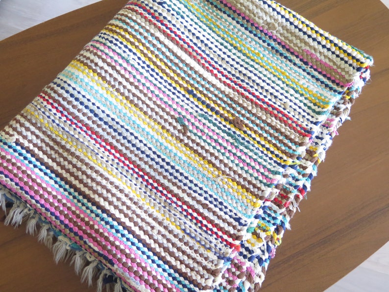 May include: A colorful striped woven rug with a fringe border. The rug is made of various colors including brown, blue, yellow, pink, and white.