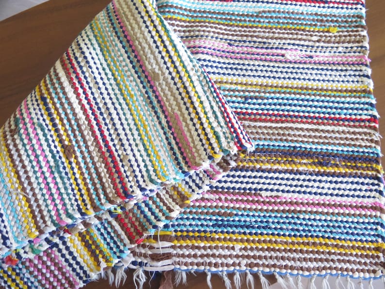 May include: A colorful woven rug with stripes of blue, yellow, pink, brown, and white. The rug is folded in half, showing the pattern on both sides.