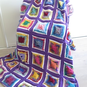 May include: A colorful crocheted throw blanket with a purple background and squares of various colors, including pink, blue, green, yellow, and red. The blanket has a textured, fluffy look.